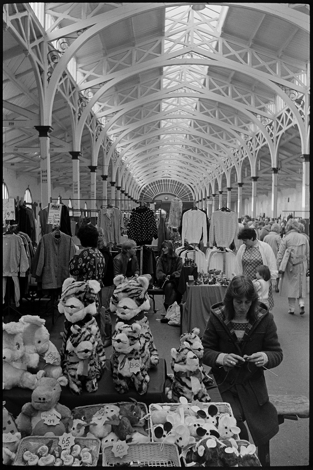 Pannier market with stalls of fruit, vegetables, eggs. 
[A woman running a stall selling soft toys, including tigers, mice and rabbits, at Barnstaple Pannier Market. She is sat by the stall knitting. Customers looking at a clothes stall can be seen in the background.]