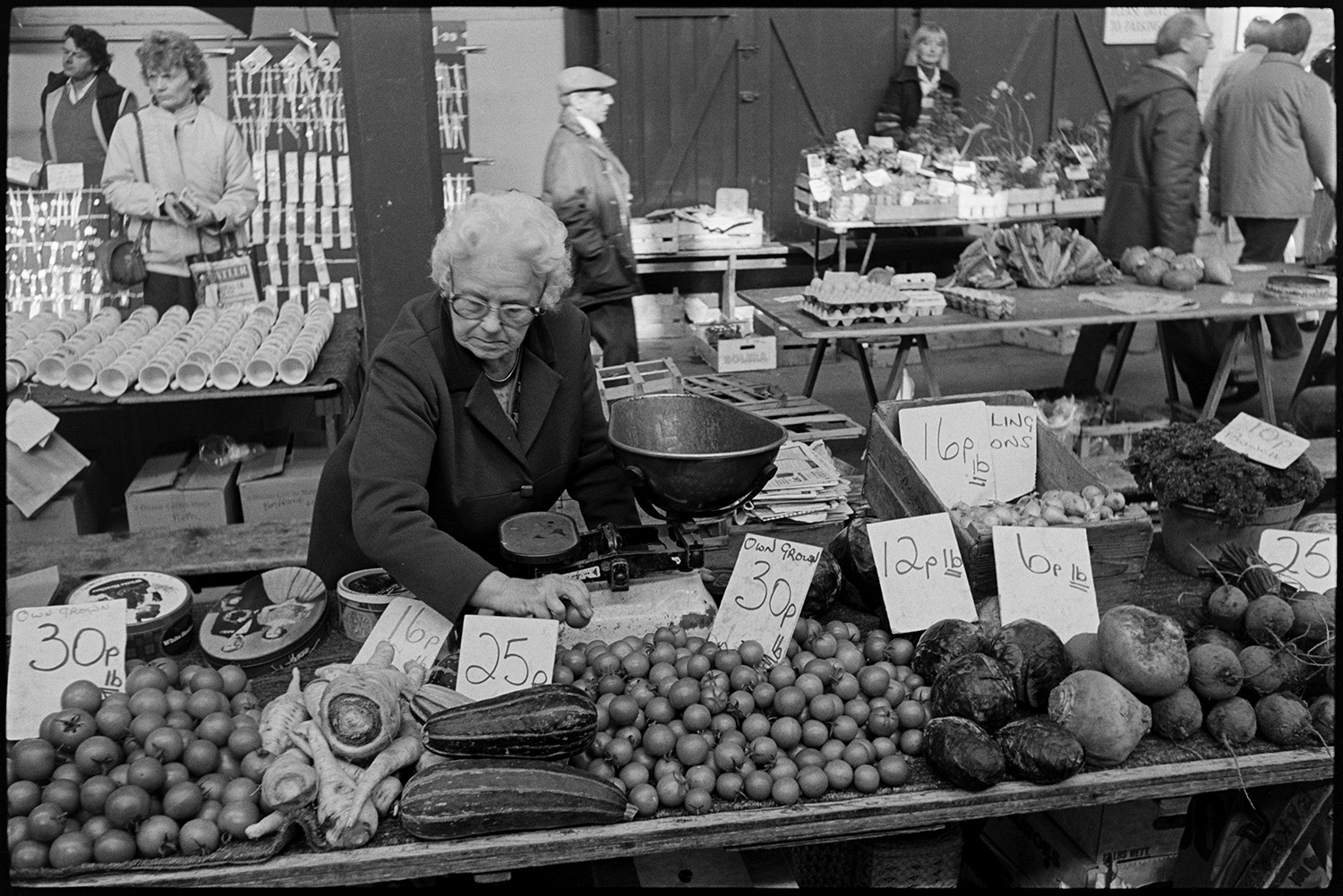 Pannier market with stalls of fruit, vegetables, eggs. 
[A woman running a vegetable stall at Barnstaple Pannier Market. She is selling tomatoes which are home grown, marrows, parsnips, swedes, beetroot and onions. There is also a set of scales on the stall. Other stalls with eggs and mugs can be seen in the background.]