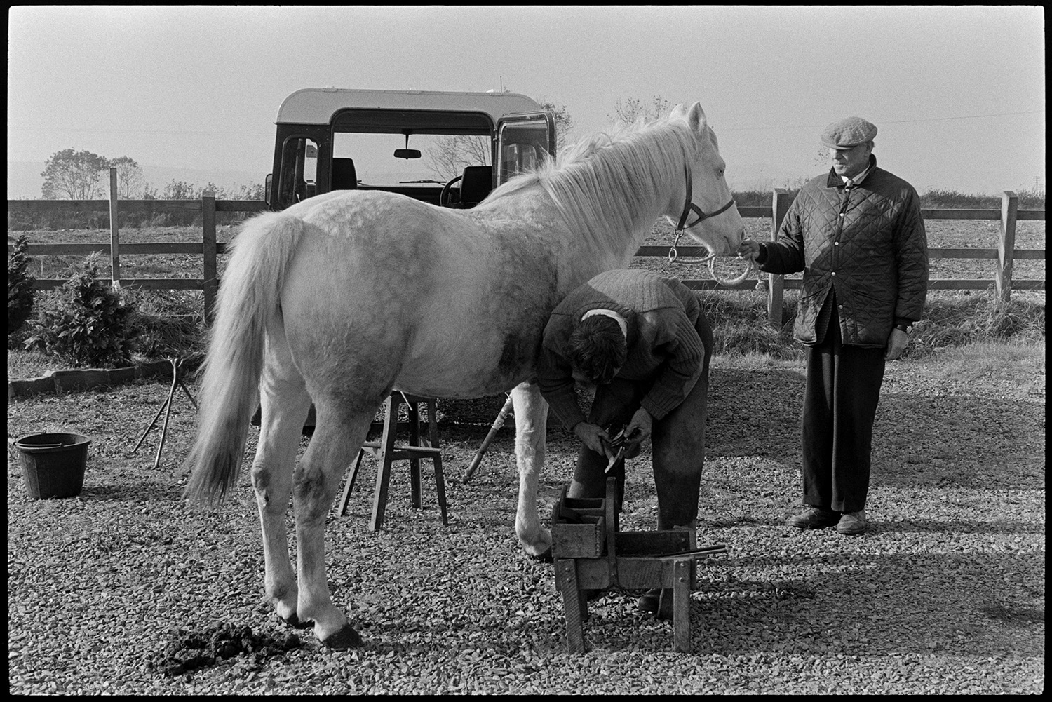 Blacksmith shoeing horse. 
[A blacksmith shoeing a horse on a gravel area outside the Beaford Centre at Greenwarren House, Beaford. Henry Bright is holding the reigns of the horse. A Land Rover is parked behind them.]