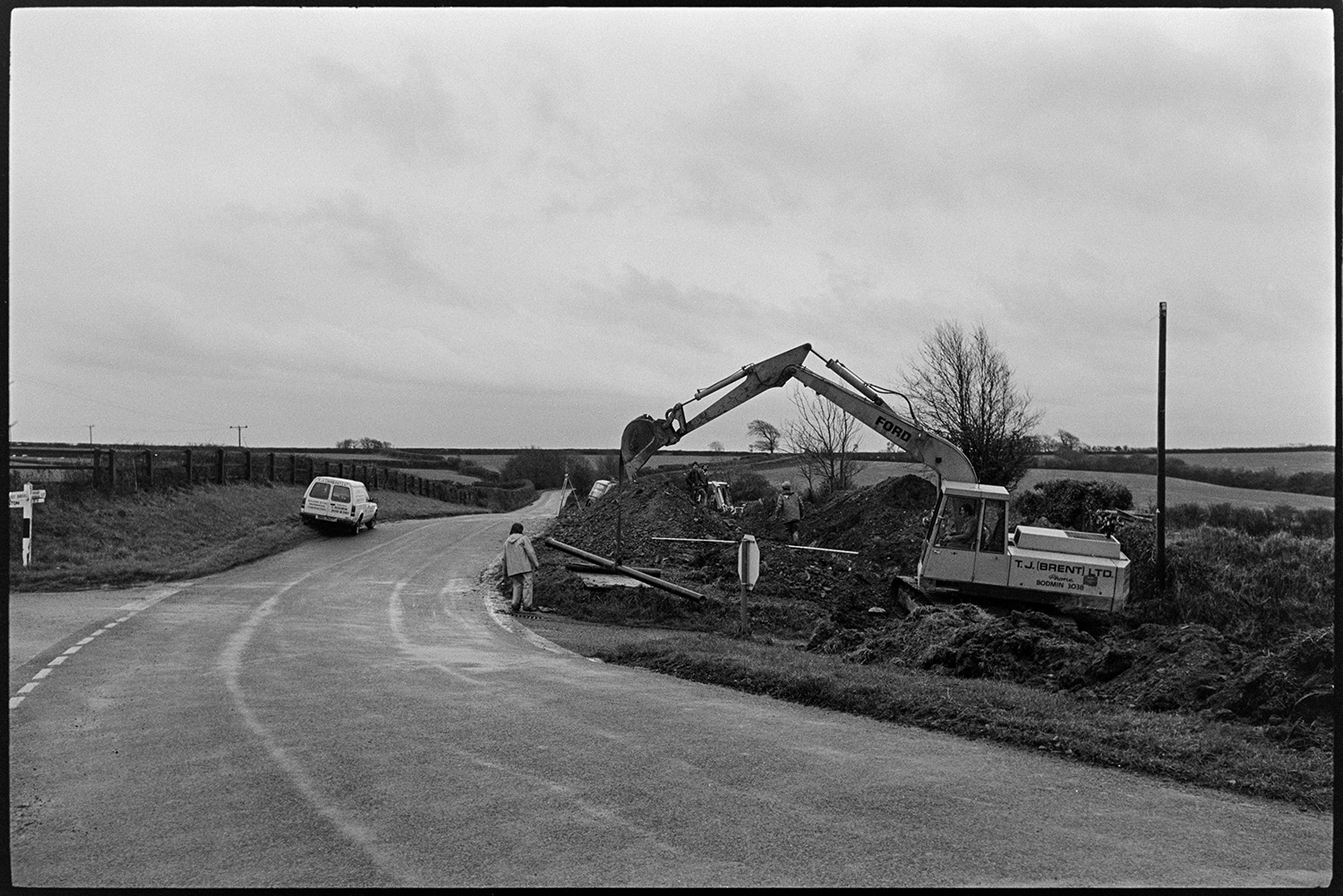 Digger clearing site of demolished house. 
[A digger clearing the site of a demolished house by a roadside at Week Cross, Burrington. Workmen are also on the site. A van is parked on the opposite side of the road near a signpost.]