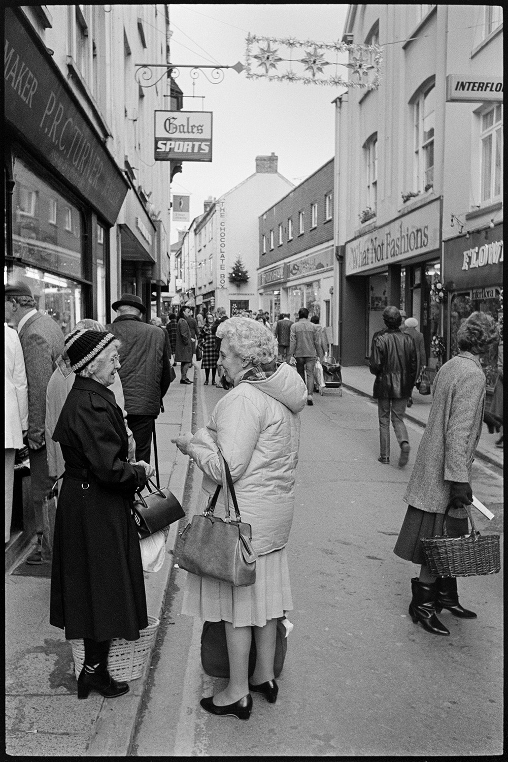 Town street scenes with women chatting holding shopping baskets. 
[Two women talking in Mill Street, Bideford. They are holding handbags and have shopping baskets with them. Other shoppers can be seen in the street as well as shop fronts, including, 'PRC Turner, Jeweller' and 'What Not Fashions'. The street has Christmas decorations hung across it.]