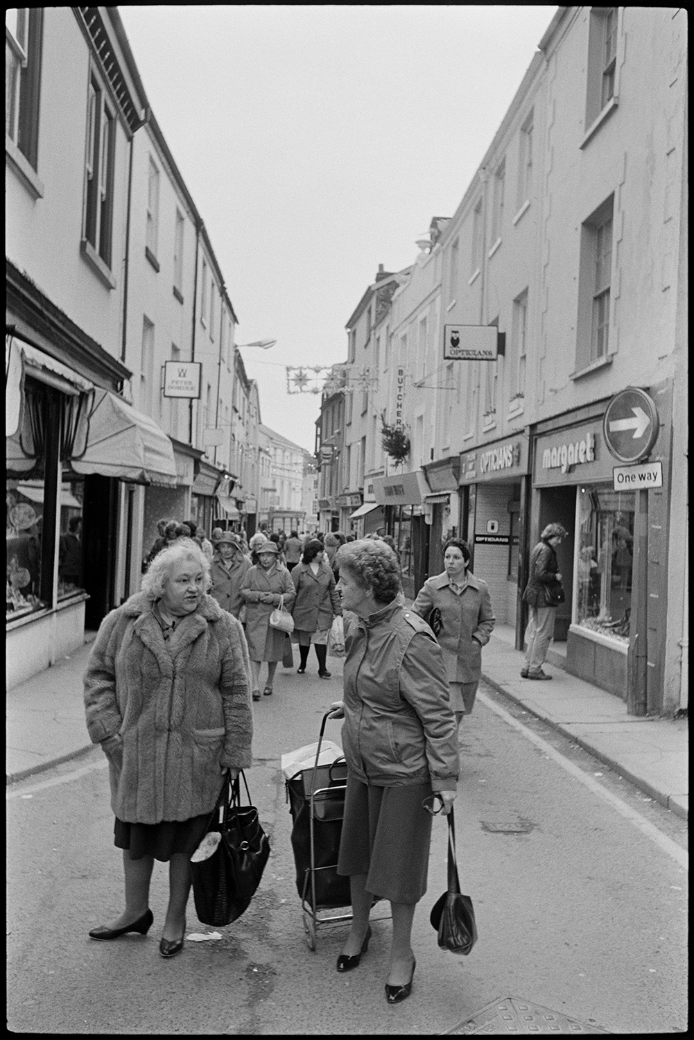 Town street scenes with women chatting holding shopping baskets. 
[Two women talking in Mill Street, Bideford. One of them is pulling a shopping basket. Other shoppers can be seen in the street as well as shop fronts, including an opticians and butcher. The street has Christmas decorations hung across it.]