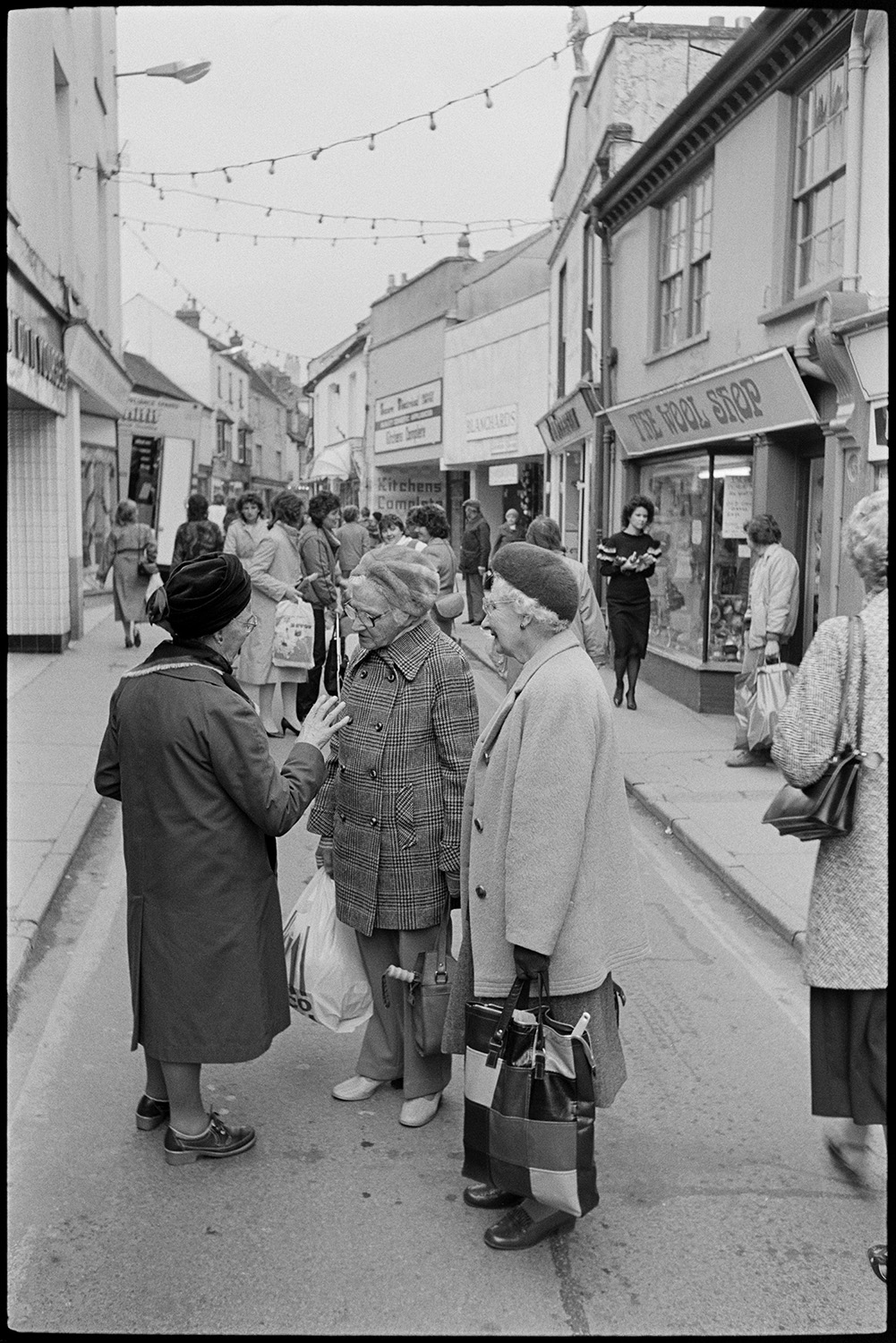 Town street scenes with women chatting holding shopping baskets. 
[Three women talking in Mill Street, Bideford. They are holding hopping bags. Other shoppers can be seen in the street as well as shop fronts , including 'The Wool Shop'. Lights are hung across the street for Christmas.]