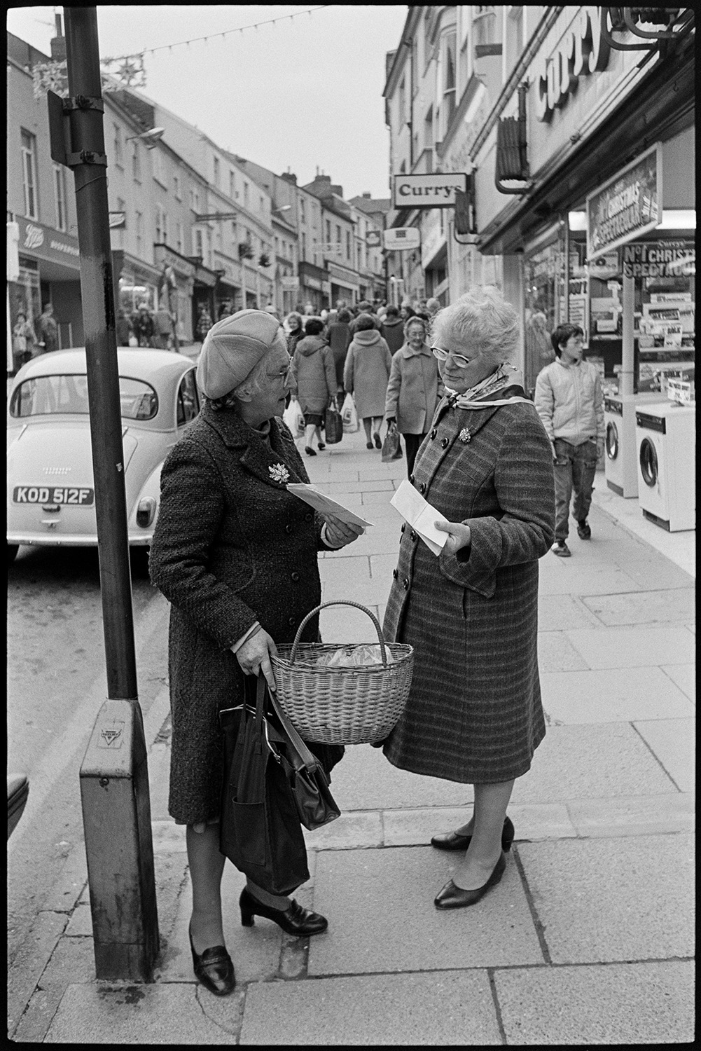Town street scenes with women chatting holding shopping baskets. 
[Two women talking in Mill Street, Bideford. One of them is holding a basket. Other shoppers can be seen as well as shop fronts, including 'Currys'.]