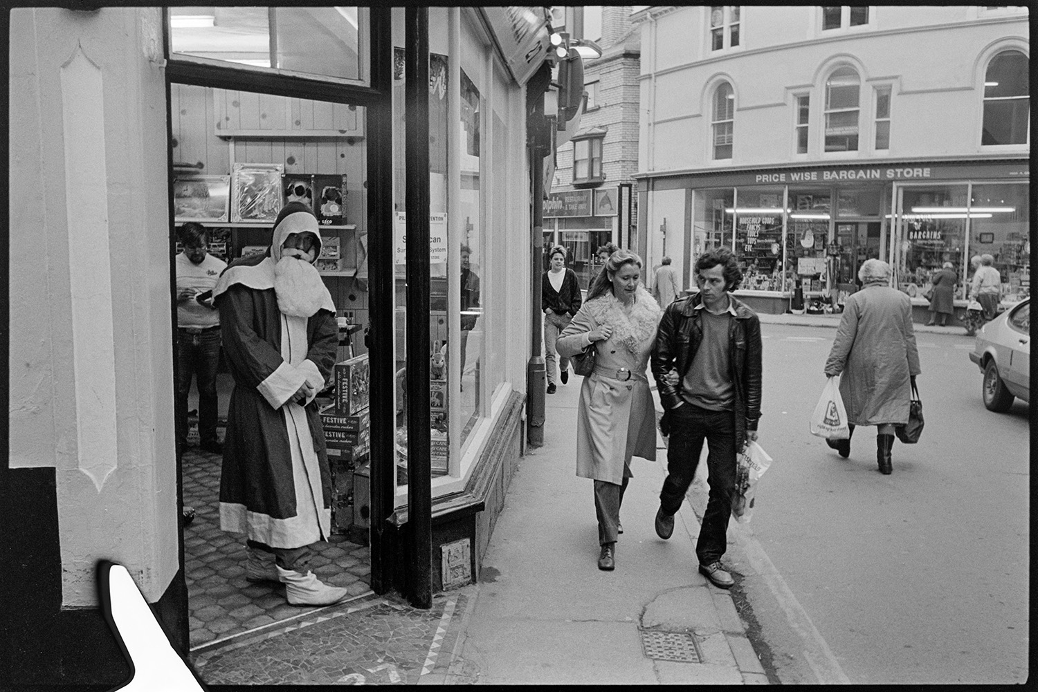 Town street scenes with women chatting holding shopping baskets. 
[A person dressed as Father Christmas in the entrance to a shop in Mill Street, Beaford. Shoppers, including a couple are walking past outside and the shop front of 'Price Wise Bargain Store' is visible in the background.]
