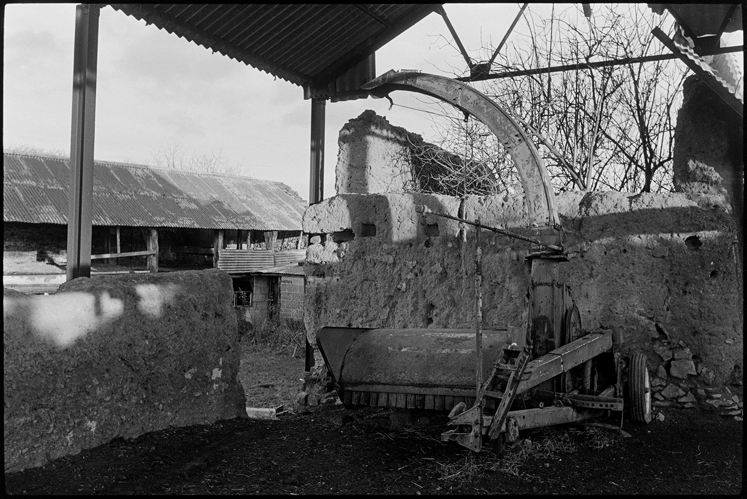 Derelict collapsing farmhouse cob and thatch old cob barn. 
[Old farm machinery in a crumbling cob and corrugated iron barn at Combe Farm, Upcott, Dolton.]