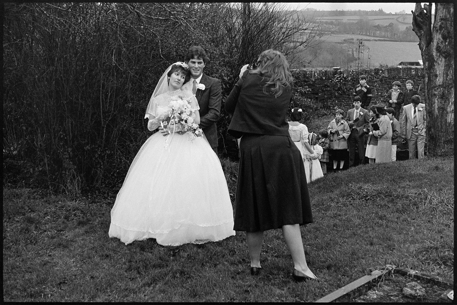 Bride, groom and guests outside church after wedding, photographer. 
[A woman photographing a bride and groom in Dolton churchyard after their wedding. Wedding guests are watching nearby.]