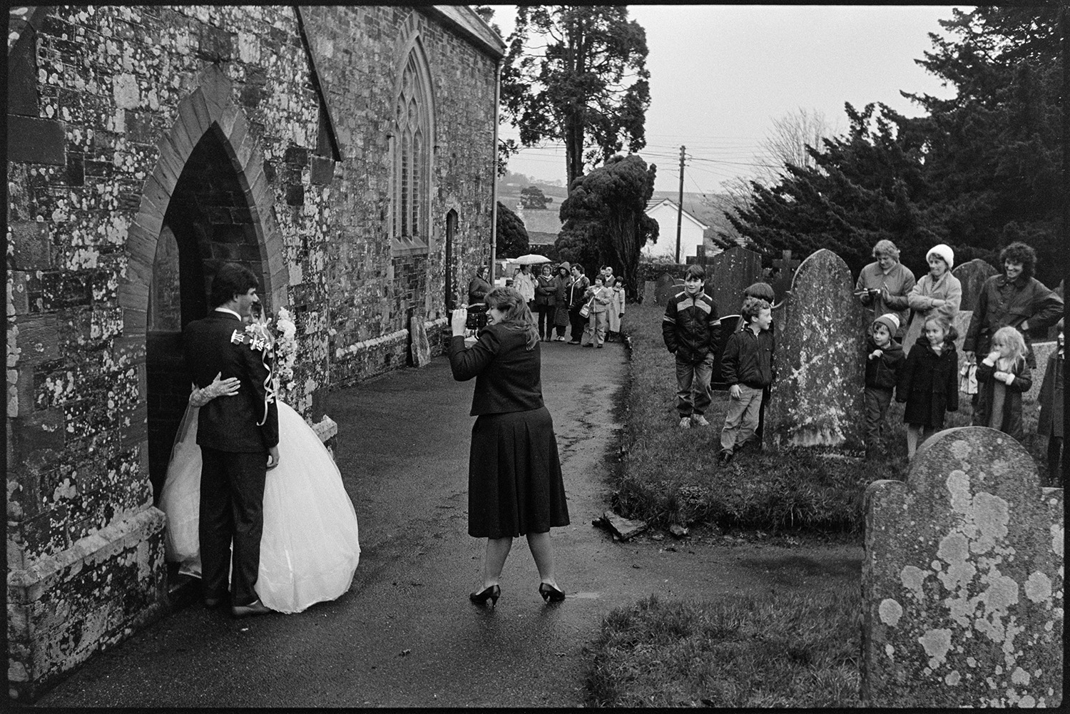 Bride, groom and guests outside church after wedding, photographer. 
[A woman photographing or videoing a bride and groom outside Dolton Church after their wedding. They are stood at the entrance to the church. Wedding guests and onlookers are watching in the background.]