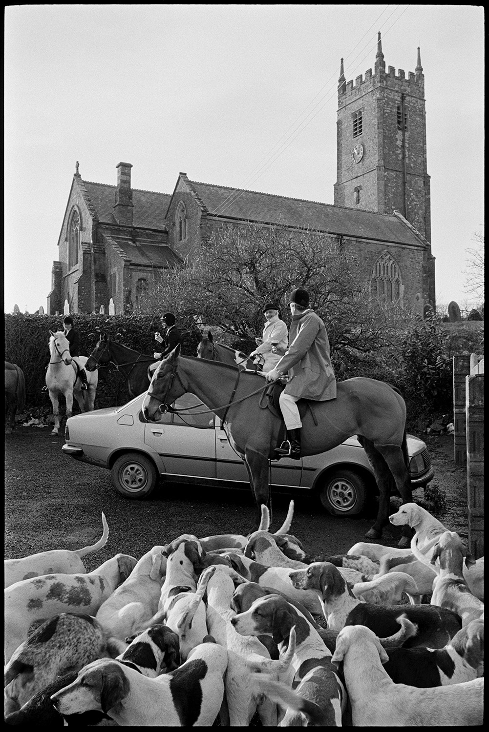 Hunt meet in village, setting off, horses and riders waiting in rain on lonely road. 
[A hunt meet by Petrockstowe Church. Huntsmen on horseback and hounds are gathered by a parked car outside the church.]