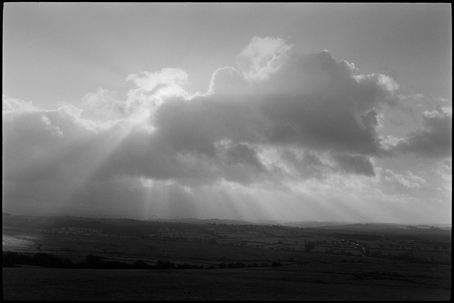 Cloudy views across moor couple walking with pram. 
[Shafts of sunlight shining through clouds onto a landscape of fields and trees on Hatherleigh Moor.]