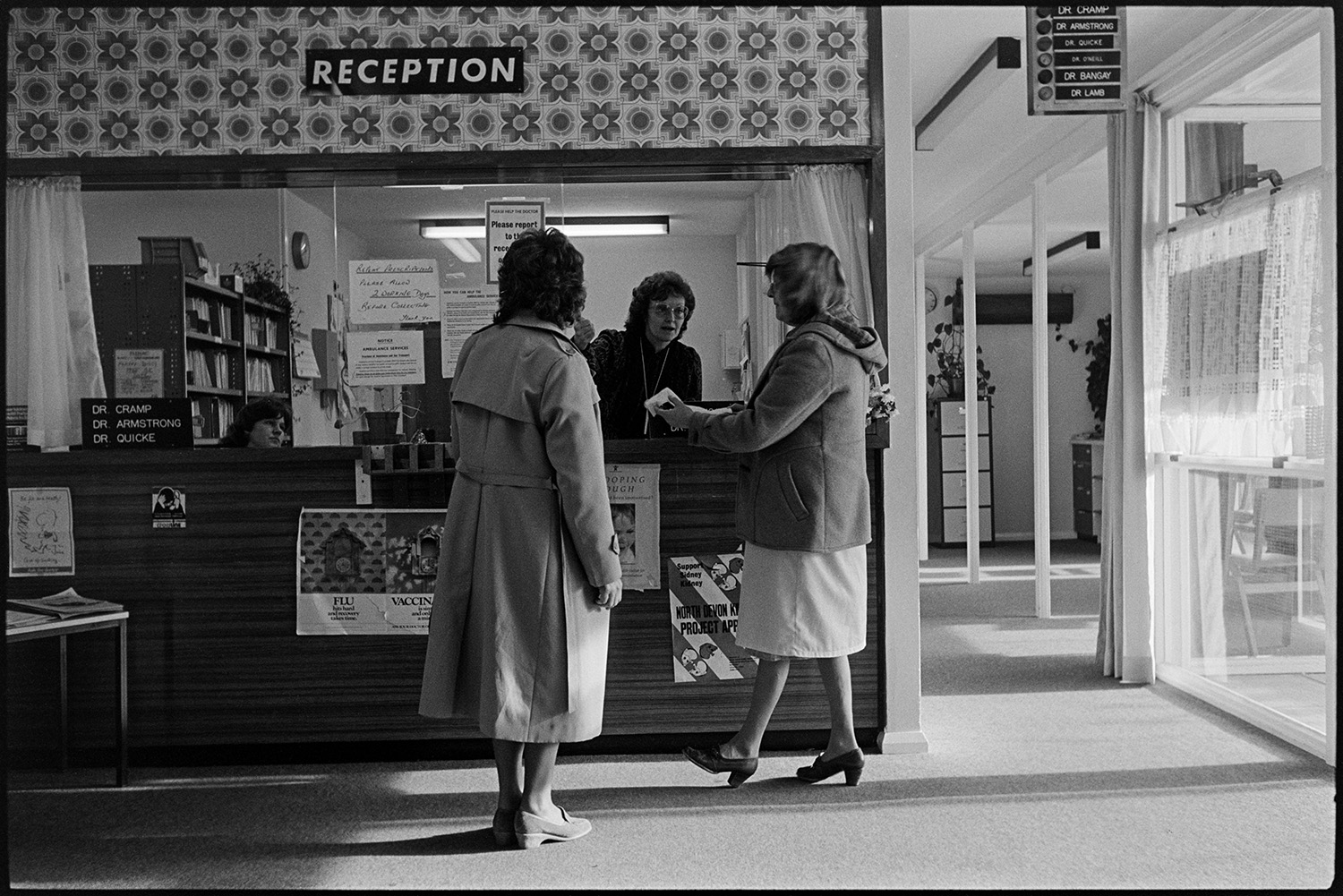 Health Centre reception desk people collecting medicine. 
[Two women collecting medicine from the reception desk at Torrington Health Centre. Various posters are displayed at the desk.]