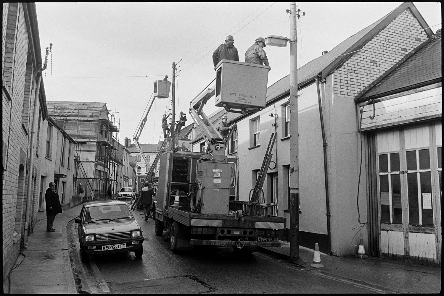 Men repairing street lights from mobile gantry. 
[Two men repairing a street light from a cherry picker, in a street in Torrington. More men are up a ladder and another cherry picker working on the telegraph pole in the background. A car is driving along the street and a building covered in scaffolding can be seen further along the road.]
