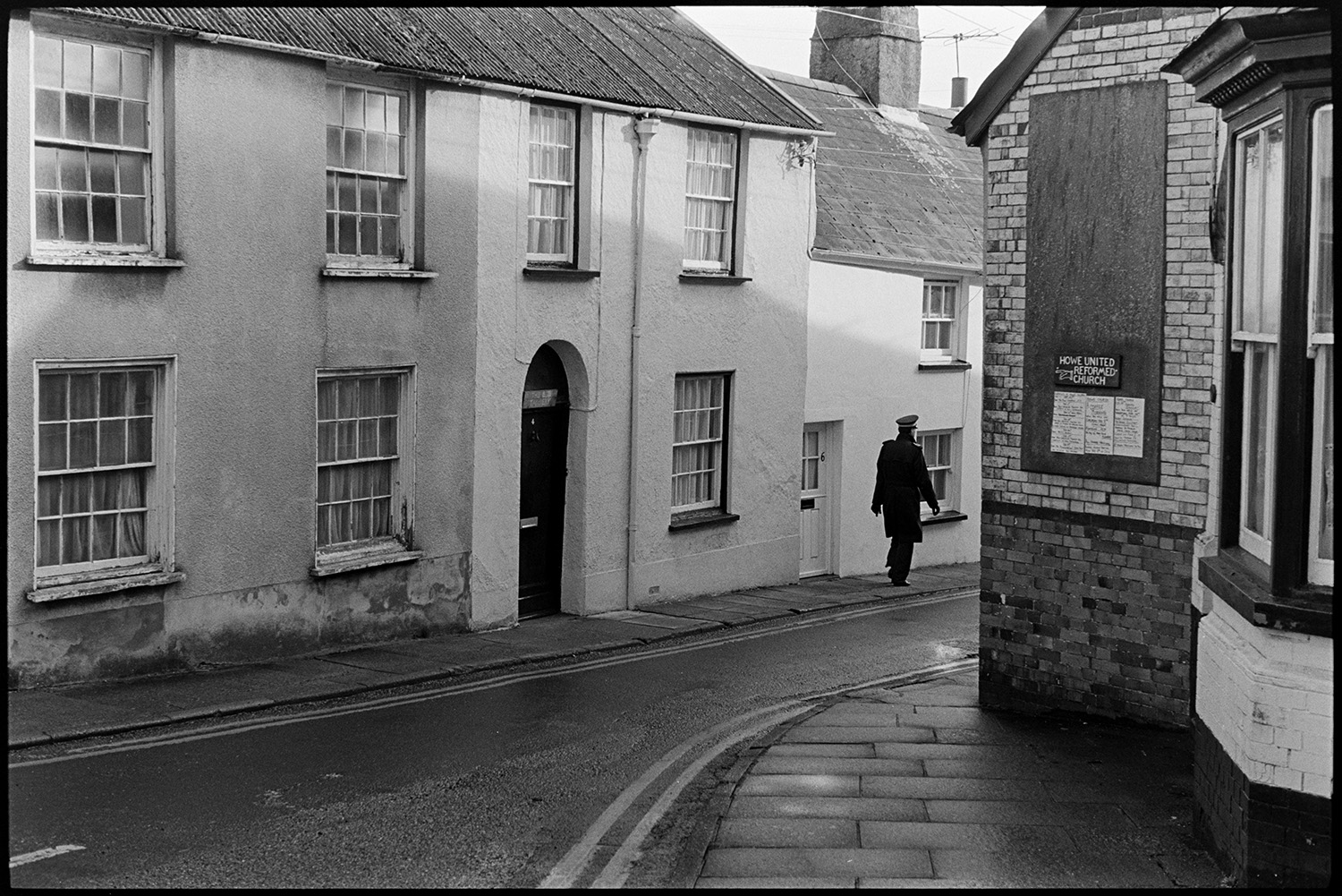 Street scene, nice windows. 
[A man in uniform, possible a policeman, walking along Castle Street in Torrington, past terraced houses. A signpost to Howe United Reformed Church is on one of the buildings in the street.]