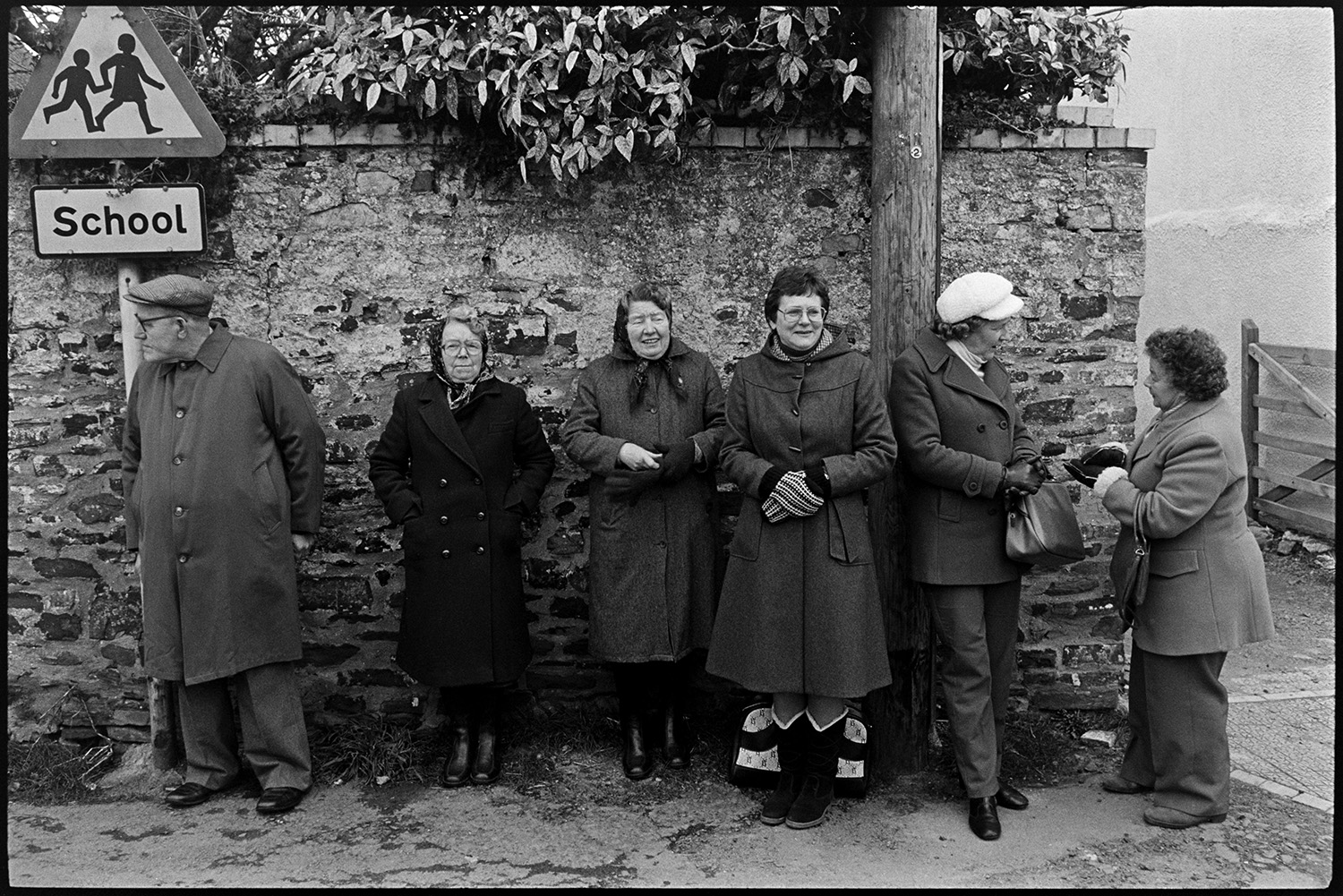 Pancake race in village street, children and spectators. 
[A man and five women stood by a stone wall in Fore Street, Dolton watching a pancake race in the street. Two of the women are talking.]