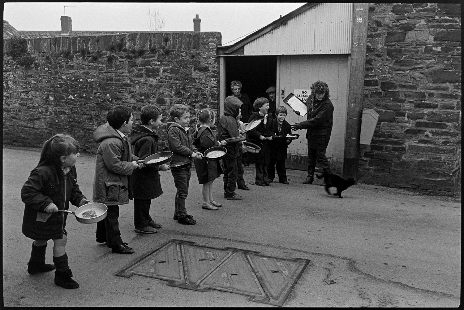 Pancake race in village street, children and spectators. 
[Children lined up holding frying pans with pancakes for a pancake race in Fore Street, Dolton. A woman holding a clip board is about to start the race as a cat is running past her.]
