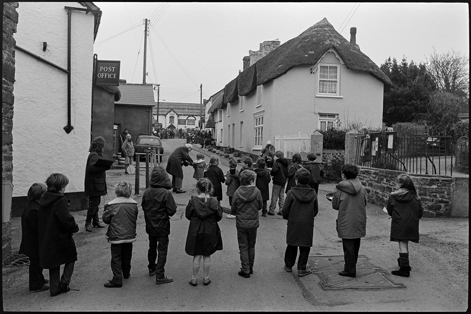 Pancake race in village street, children and spectators. 
[Children lined up in two rows with frying pans ready for a pancake race in Fore Street, Dolton. Spectators are watching from the other end of the street by the Royal Oak pub. Thatched cottages and a Post Office sign can be seen along the street.]