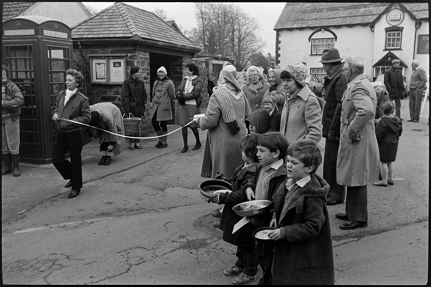 Pancake race in village street, children and spectators. 
[Three boys holding frying pans after taking part in a pancake race in Fore Street, Dolton. Men and women are watching the races outside the Royal Oak pub and two women are holding a finishing line by a telephone box and bus shelter.]