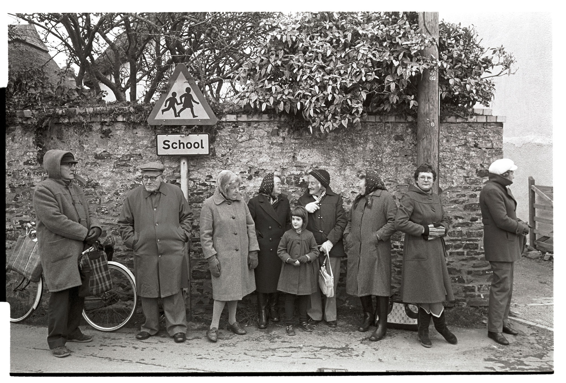 Pancake race in village street spectators standing against wall at finish.
[A group of people and a child, Ella Ravilious, standing against a wall in Fore Street, Dolton,  on Shrove Tuesday, waiting for the finish of the village pancake race. They are wearing coats against the cold and one man is stood by a bicycle.]