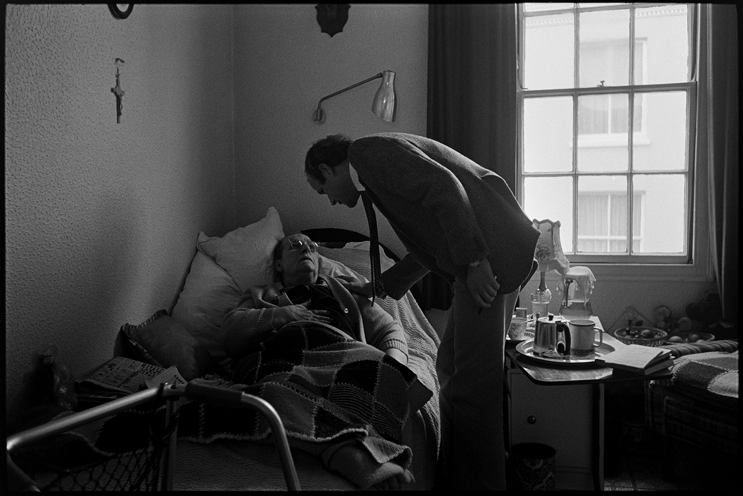 Doctor on his rounds talking woman patient in bed in cottage hospital. 
[Doctor Richard Westcott visiting a patient in South Molton Cottage Hospital. A bedside table with a lamp, mug and teapot is visible.]
