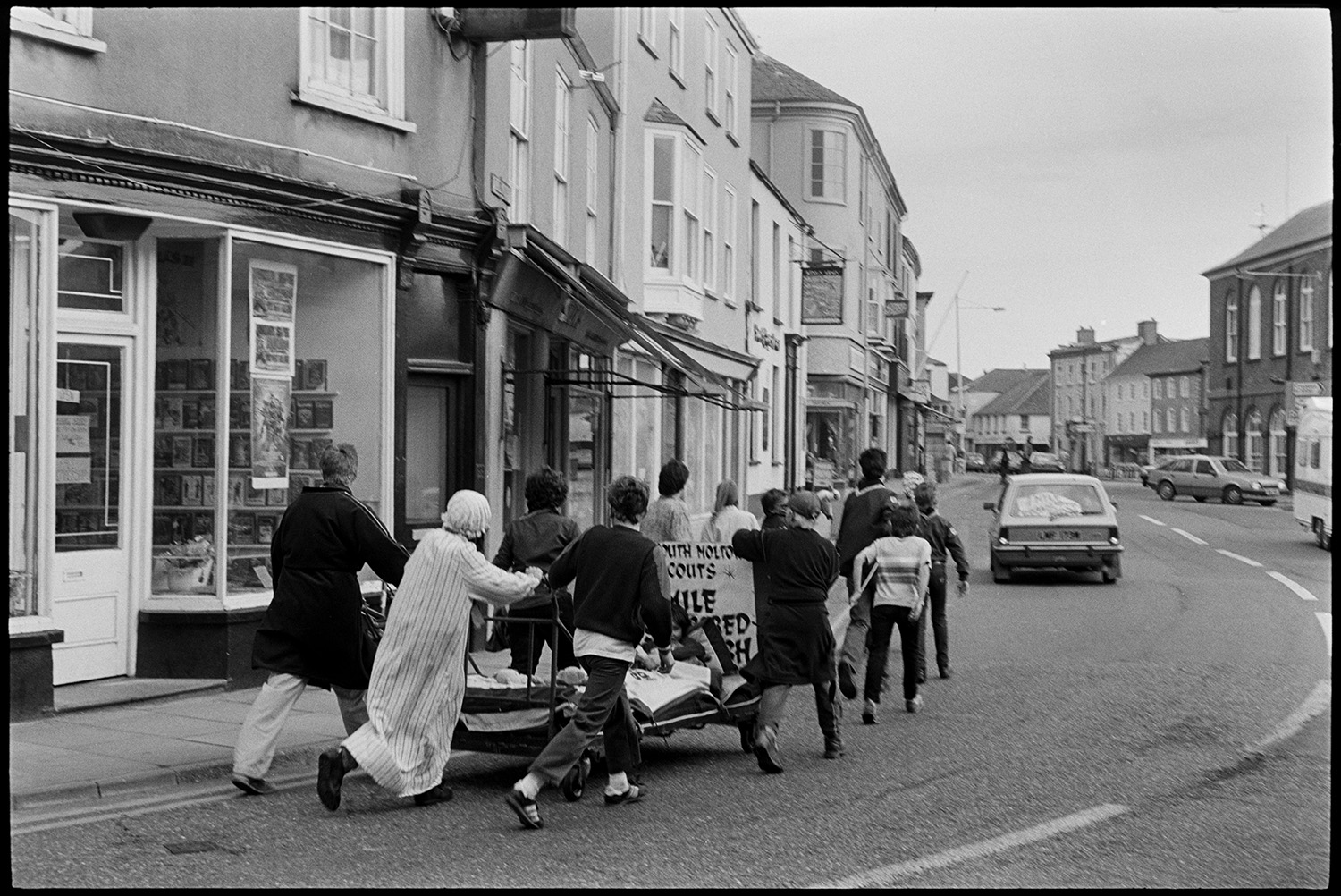 Scouts on sponsored walk pushing bed through town. 
[A group of Scouts on a 7 mile sponsored walk pushing and pulling a make-shift bed on wheels, with a child in it, through a street in South Molton, past shop fronts. Some of them are in fancy dress. A car is driving ahead of them.]