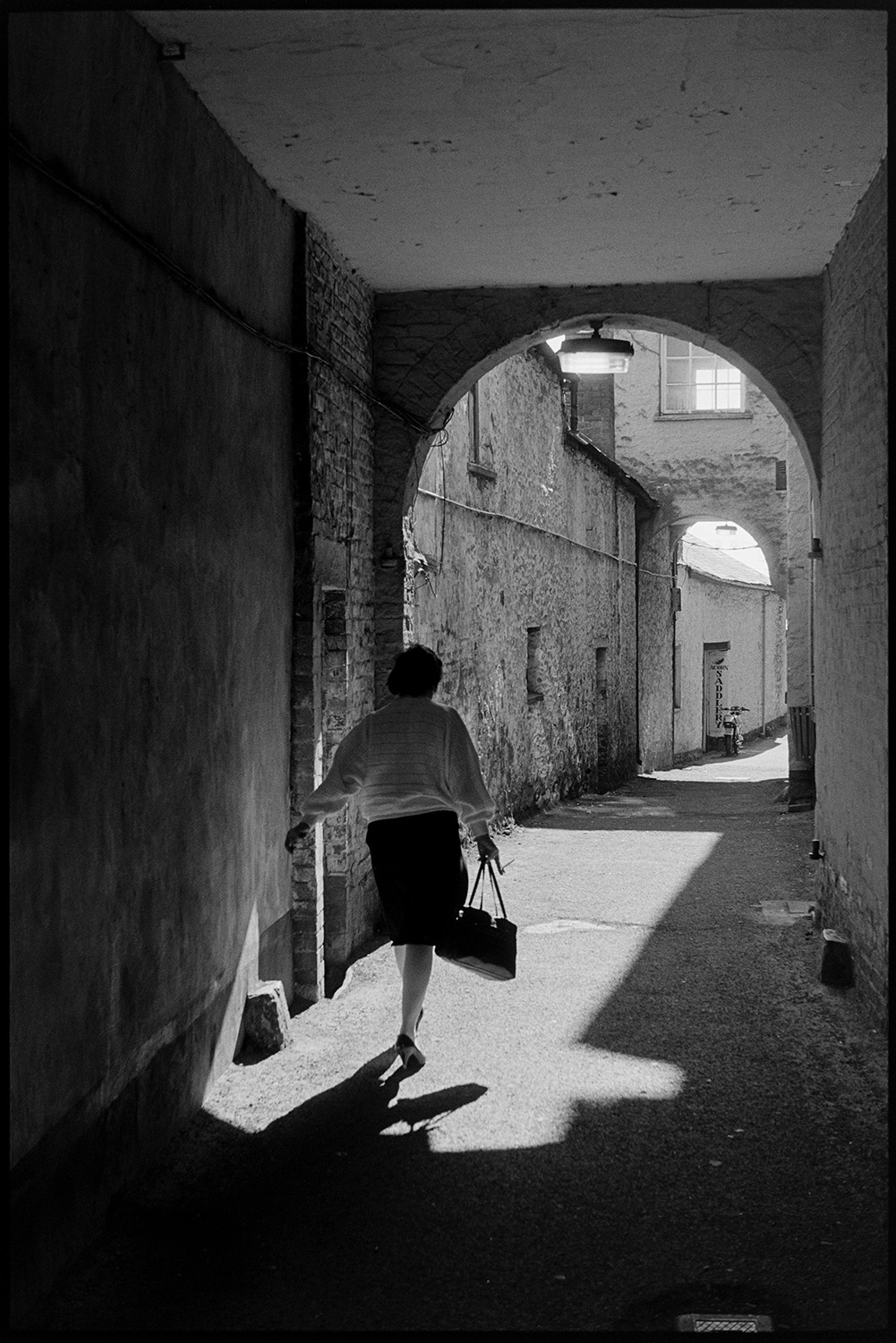 Covered alleyway with passers by, sunlight. 
[A woman carrying a bag walking through an arched alleyway by South Molton Town Hall. Sunlight is shining into the alleyway.]