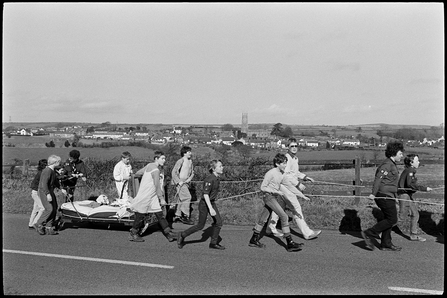 Scouts on sponsored walk pushing bed along country road to South Molton. 
[A group of Scouts on a 7 mile sponsored walk pushing and pulling a make-shift bed on wheels, along a road near Chittlehampton, with the town in the background. Some of the children are in fancy dress. Chittlehampton Church can be seen in the background.]