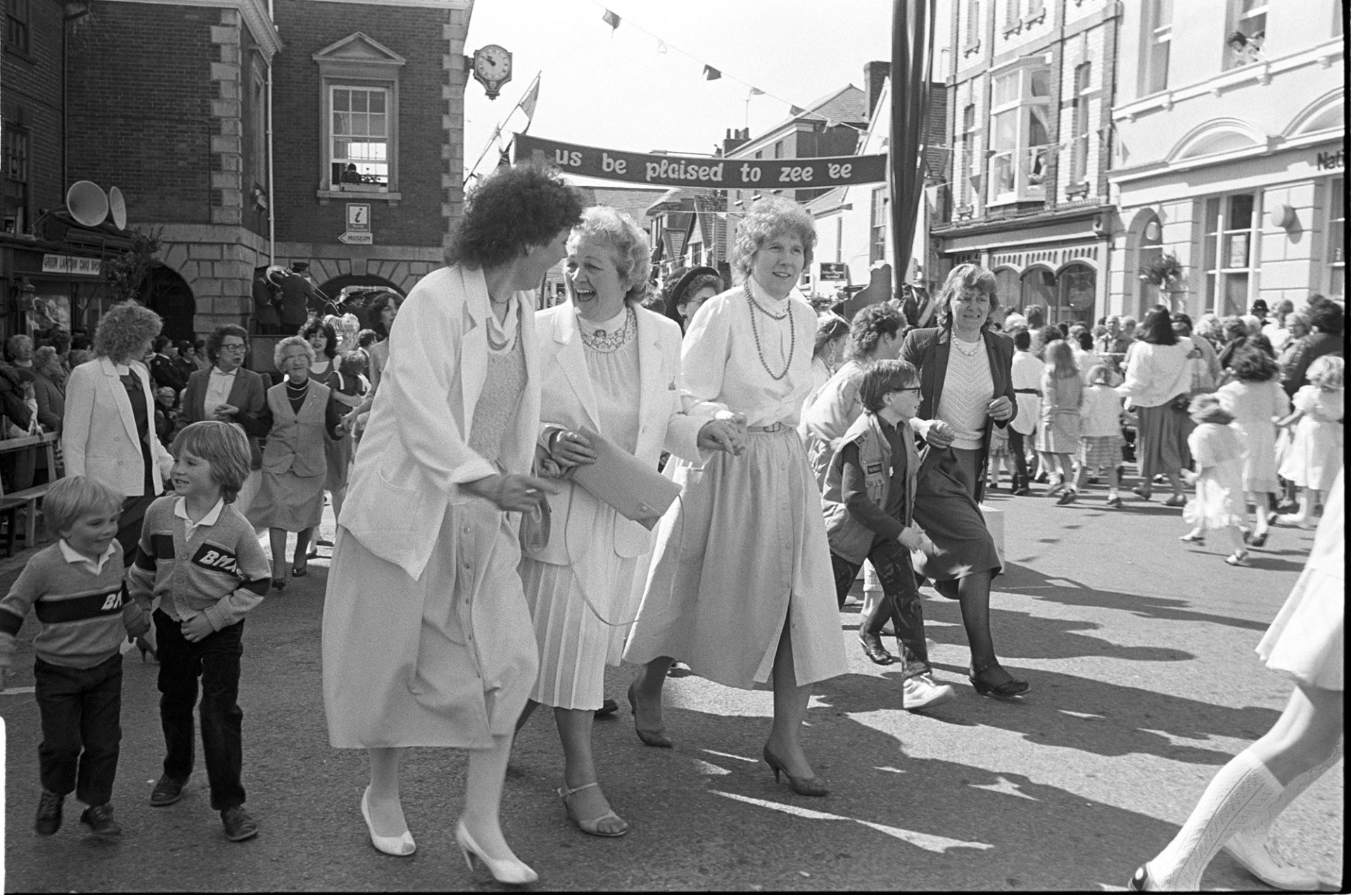 May Fair celebrations, dancing in the street, three women in summer dresses.
[Three women in summer clothes laughing and dancing in the High Street at the Great Torrington May Fair celebration. Stretched across the street is a banner reading 'Us be plaised to zee 'ee'. Other people are in the street watching the celebrations.]