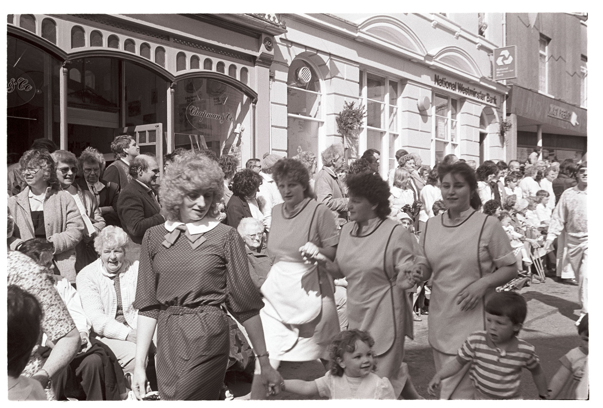 May Fair celebrations, dancing in the street, three women in working overalls.
[Three women in overalls or aprons dancing in the High Street at the May Fair celebrations in Great Torrington. In front of them is a woman with three small children. Spectators are watching the May Fair parade from the side of the street outside shop fronts.]