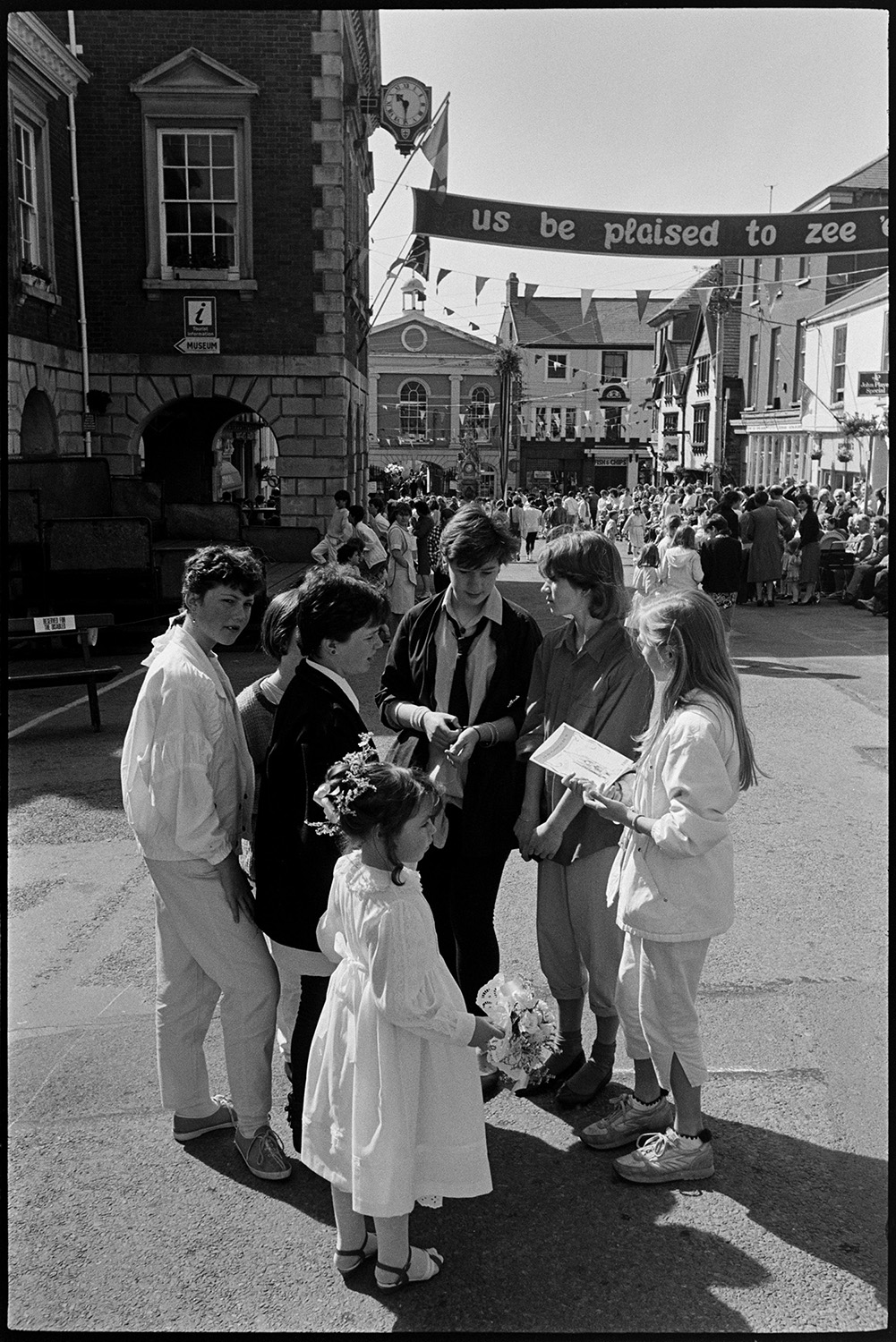 May Fair celebrations, people waiting for start of fair, preparing float. 
[A group of children talking by a young girl wearing a white dress and holding a posie of flowers, possibly one of the May Fair Queen attendants at Torrington May Fair. The Town Hall and square, which is decorated with bunting, can be seen in the background. People are gathered in the square and a banner is hung across the street.]