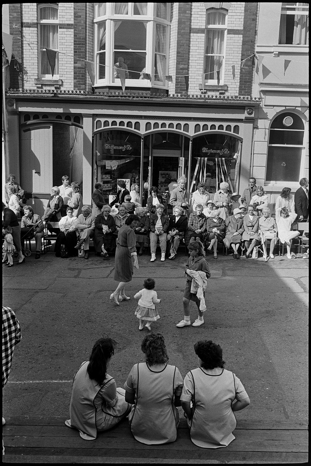 May Fair celebrations, people waiting for start of fair, preparing float. 
[A woman and two young girls dancing in a street at Torrington May Fair. People are sat on the side of the street watching, outside the shop front of Chapman & Co.]