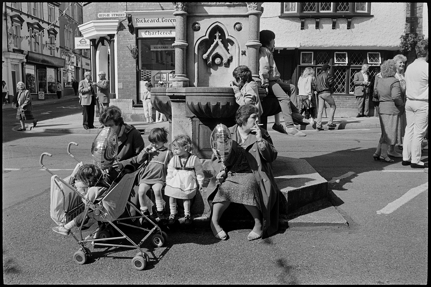May Fair celebrations, people waiting for start of fair, preparing float. <br /> [Two women and children sat by the fountain in Torrington Square waiting for the May Fair to start. One child is in a pram and one of the women is holding a balloon and smoking a cigarette. Shop fronts can be seen in the background.]