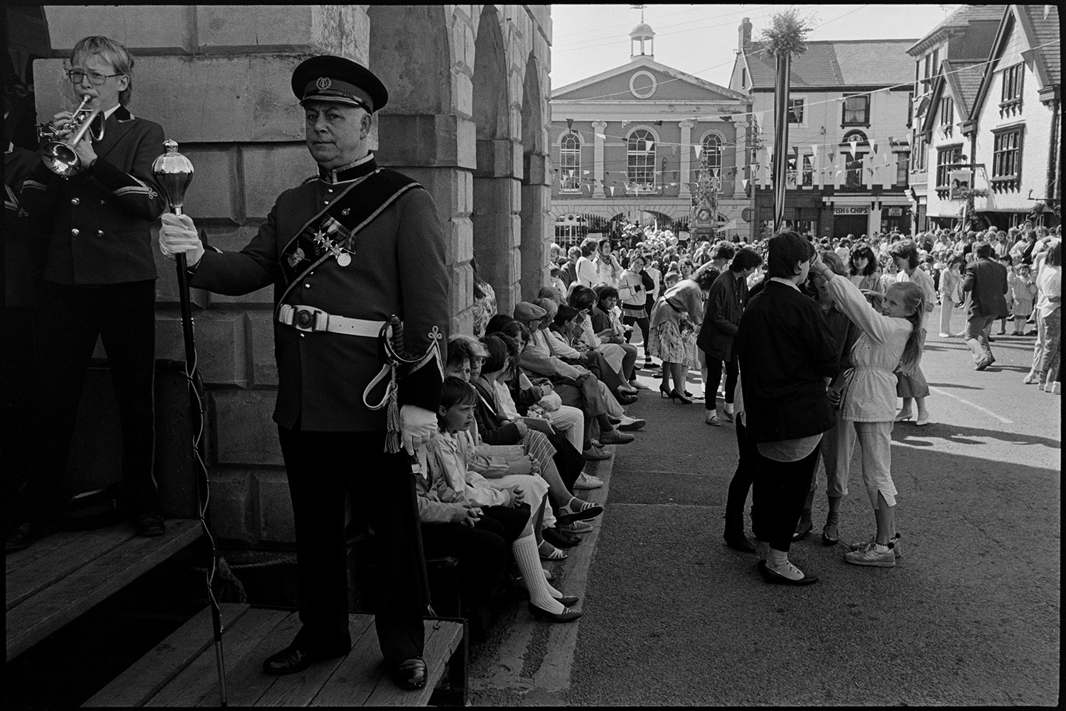 May Fair celebrations, band marching into square, children dancing. 
[People sat outside the Town Hall at Torrington May Fair, A man wearing a uniform and holding a large mace or baton is stood next to a bandsman playing a cornet. Men, women and children are dancing in the street in the background, which is decorated with bunting.]