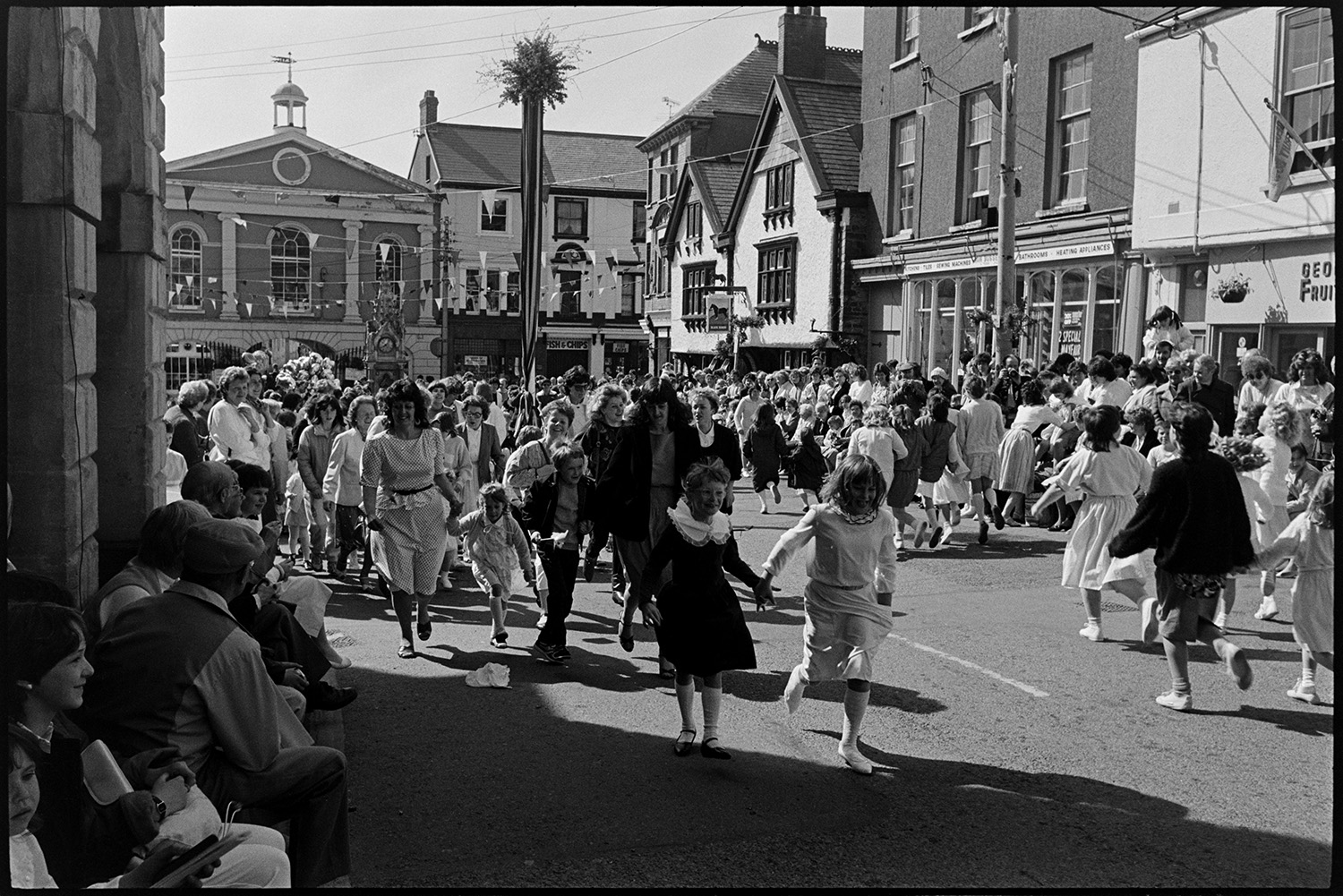 May Fair celebrations, band marching into square, children dancing. 
[Women and children dancing in the Square at Torrington May Fair. People are sat watching outside the Town Hall. The square is decorated with bunting and a maypole is visible.]
