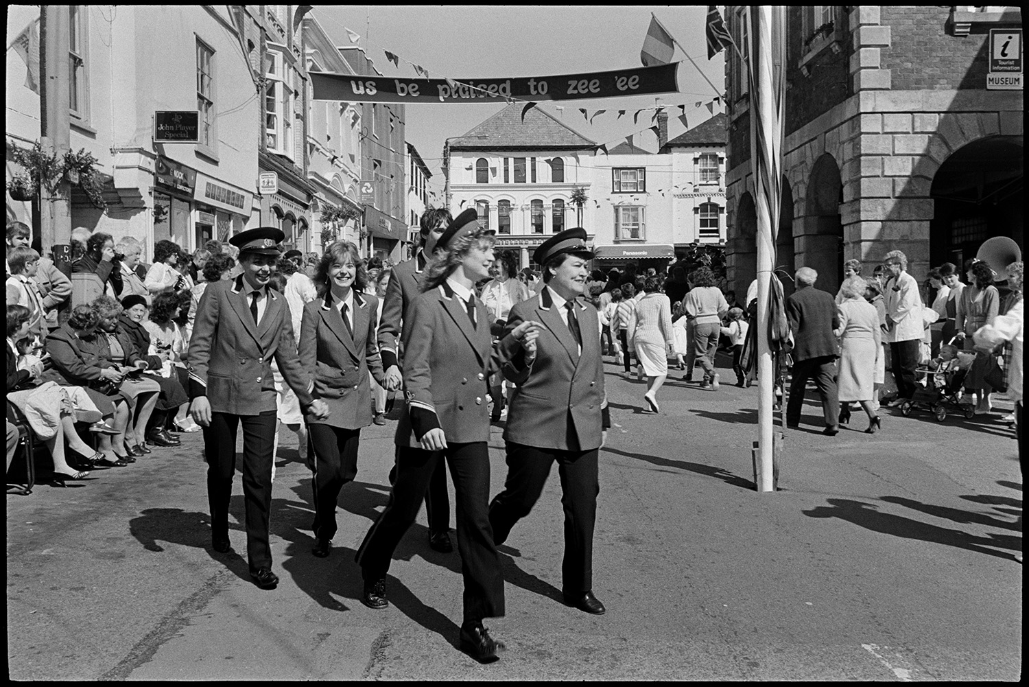 May Fair. Dancing with band marching, spectators and passers by. Maypole. 
[Women dressed in a band uniform dancing in a street outside the Town Hall at Torrington May Fair. Other people are also dancing. Spectators are watching from the side of the street and a maypole is visible in the foreground. The street is decorated with a banner an bunting.]