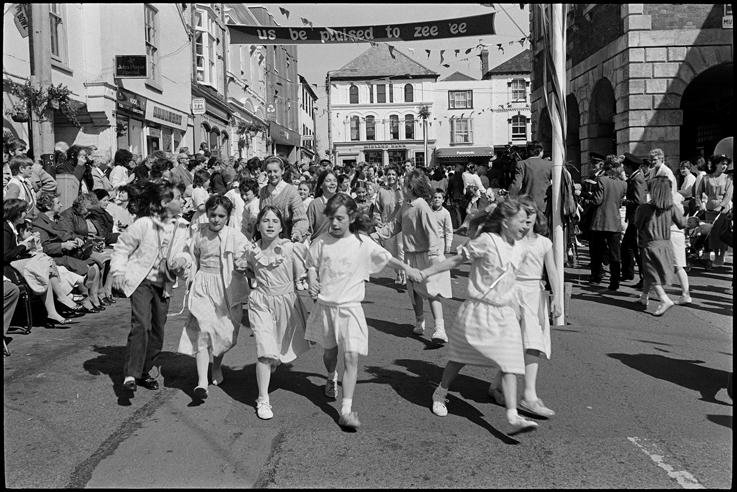 May Fair. Dancing with band marching, spectators and passers by. Maypole. 
[A group of young girls holding hands in a line dancing in a street outside the Town Hall at Torrington May Fair. Other people are also dancing. Spectators are watching from the side of the street and a maypole is visible in the foreground. The street is decorated with a banner an bunting.]
