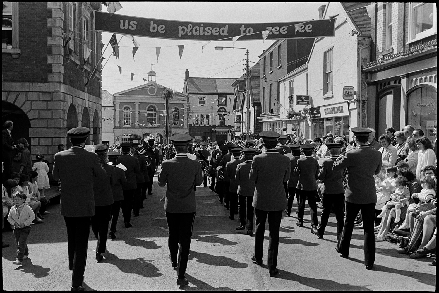 May Fair. Dancing with band marching, spectators and passers by. Maypole. 
[A marching band parading past Torrington Town Hall at Torrington May Fair. Spectators are watching from the side of the street outside shop fronts. The street is decorated with a banner and bunting.]