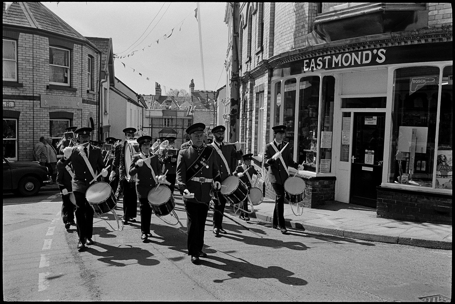 May Fair. Dancing with band marching, spectators and passers by. Maypole. 
[A marching band with drums and brass instruments parading past Eastmond's Hardware shop front at the Torrington May Fair. The street is decorated with bunting.]