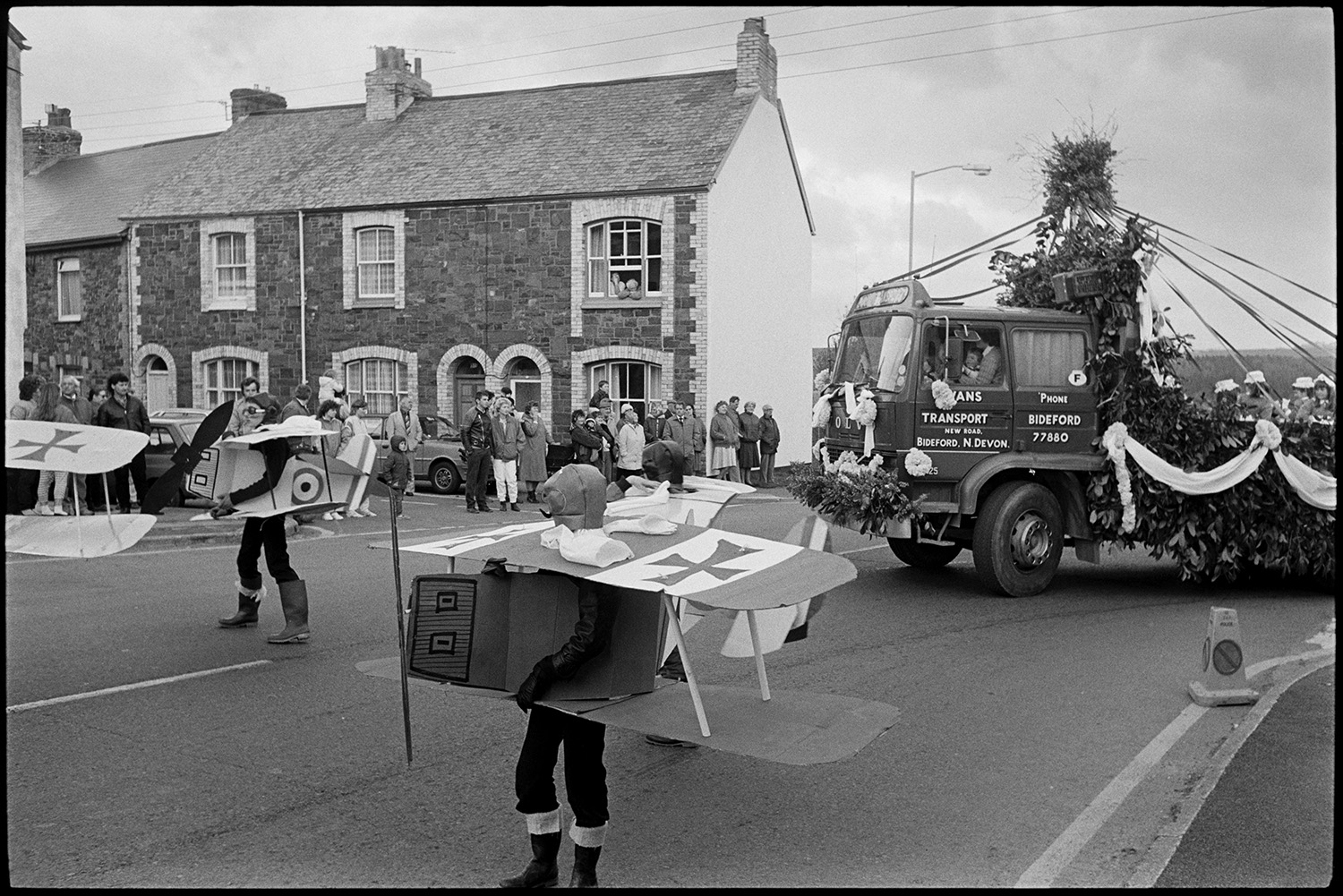 May Fair. Fancy dress parade, decorated bicycles, floats, old bicycles, penny farthing. 
[Spectators watching Torrington May Fair parade along a street. Some people in the parade are dressed as aeroplanes. They are followed by a lorry decorated with ribbons and foliage.]