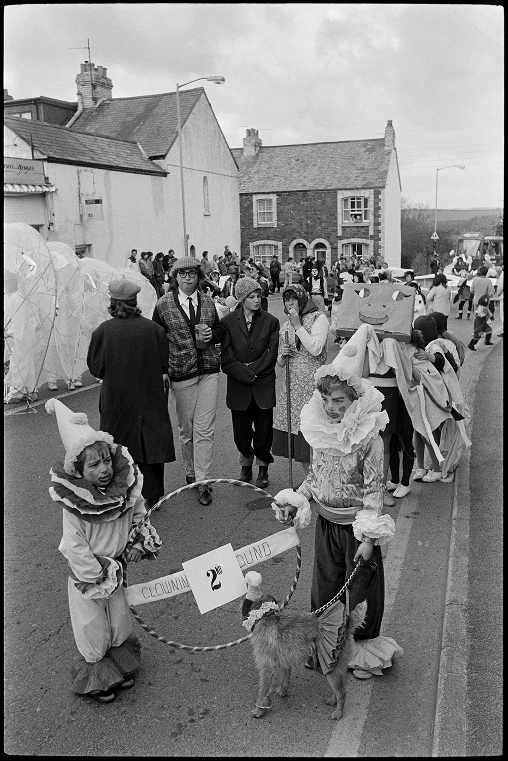 May Fair. Fancy dress parade, decorated bicycles, floats, old bicycles, penny farthing. 
[Two children dressed as clowns with a dog at Torrington May Fair parade. They are holding a hula hoop between them with a sign reading 'Clowning Around'. Other people at the May Fair, some in fancy dress, can be seen in the background.]