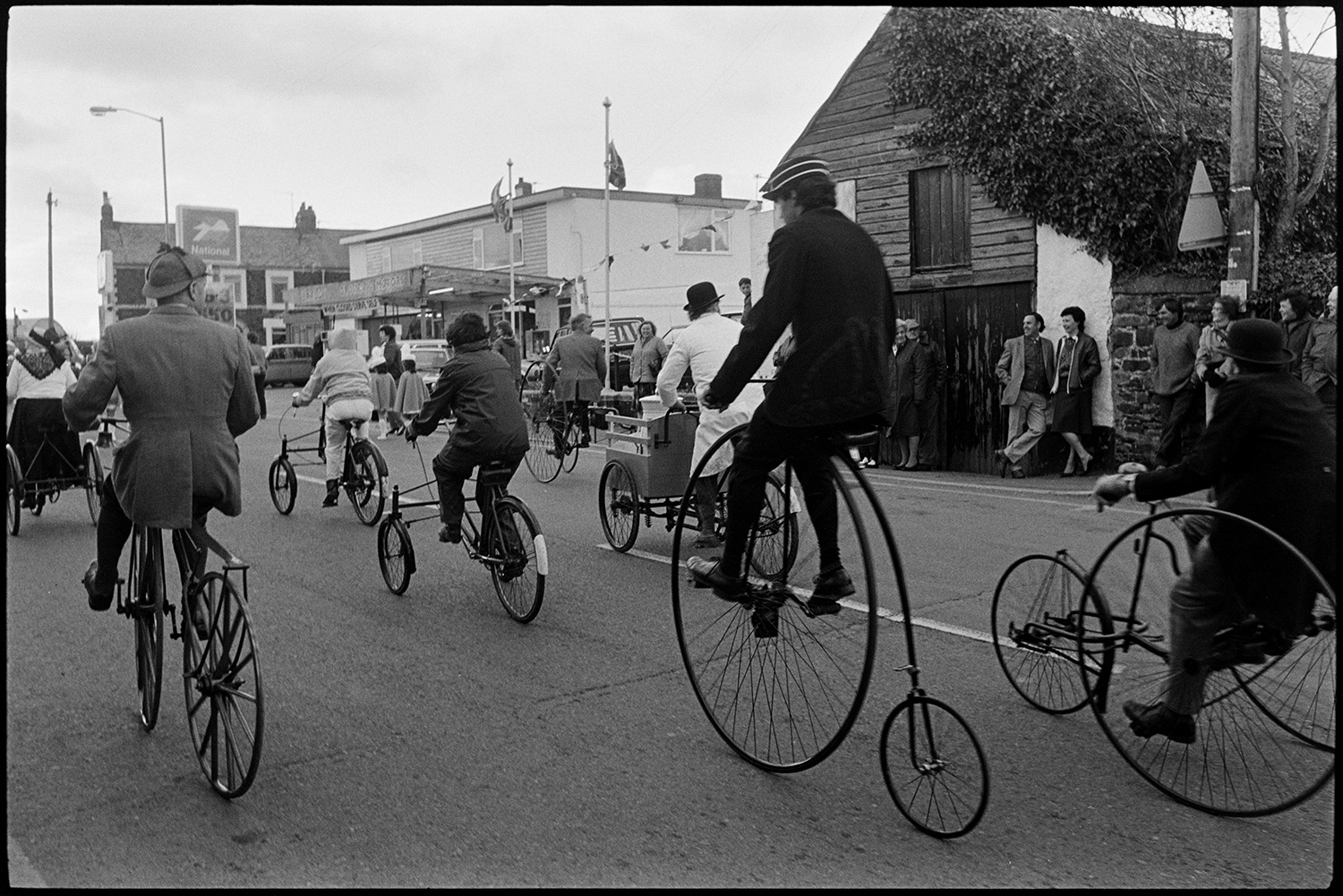 May Fair. Fancy dress parade, decorated bicycles, floats, old bicycles, penny farthing. 
[A group of people riding old bicycles, including penny farthings, in the Torrington May Fair parade. People are watching the parade from the side of the street.]
