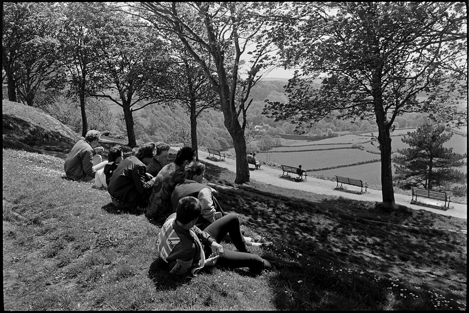 Boys in park just after leaving school. 
[A group of teenage boys sat in Castle Hill Park at Torrington, overlooking a landscape of trees, hedgerows, fields and a footpath with benches. One of them is wearing a jacket with a union jack motif.]