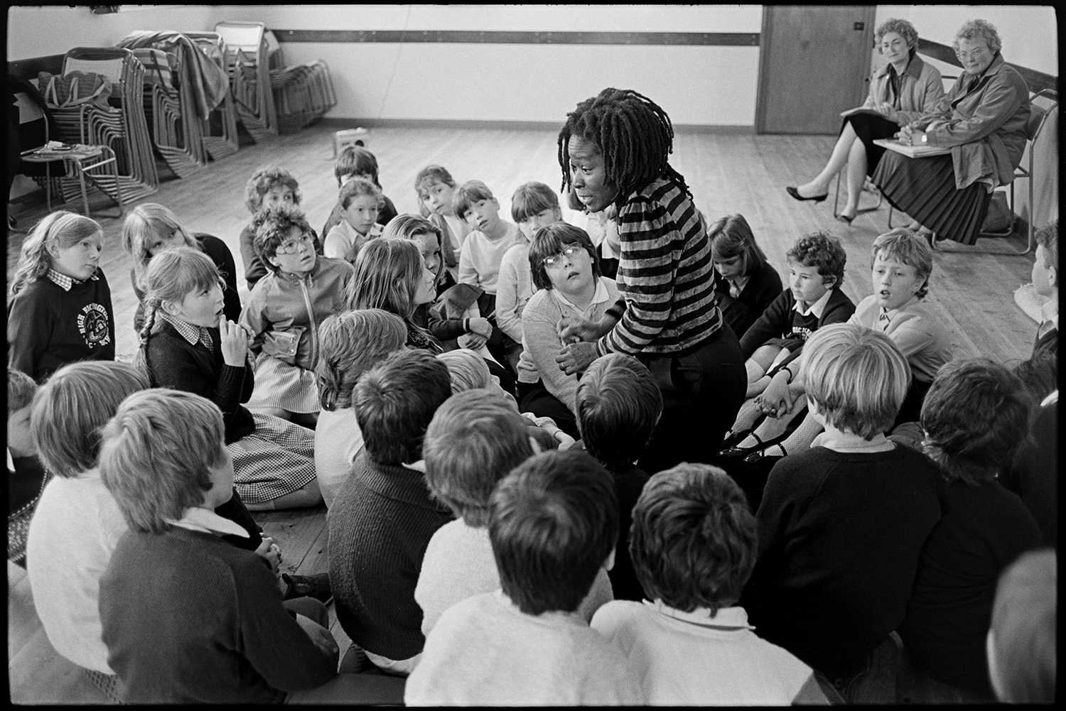 Caribbean woman writer in primary school telling stories to children. 
[Pearl Springer, a Caribbean writer, telling a story to a group of schoolchildren sat on the floor of a school hall in High Bickington. Two women are sat on chairs behind them.]