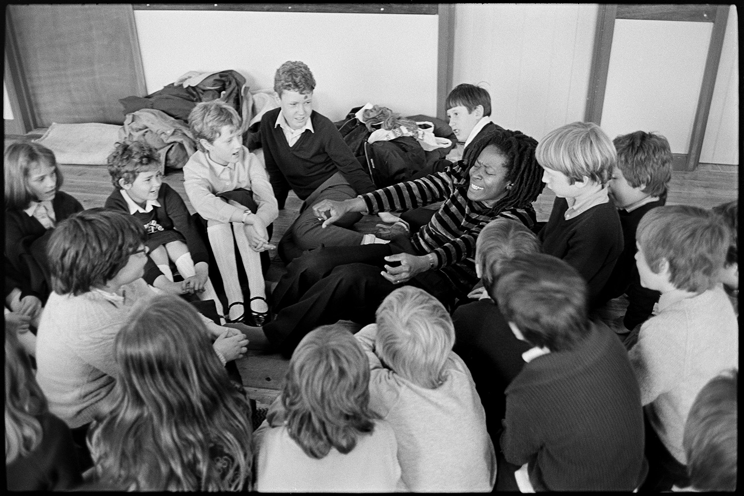 Caribbean woman writer in primary school telling stories to children. 
[Pearl Springer, a Caribbean writer, telling a story to a group of schoolchildren sat on the floor of a school hall in High Bickington. She is acting part of the story.]