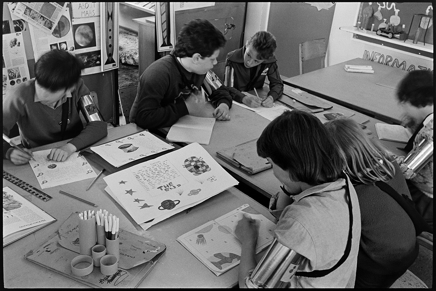 Schoolchildren working on space project pretending to be astronauts. 
[Boys and girls drawing designs for a space project in a classroom at Bideford Primary School. They are all wearing arm bands.]