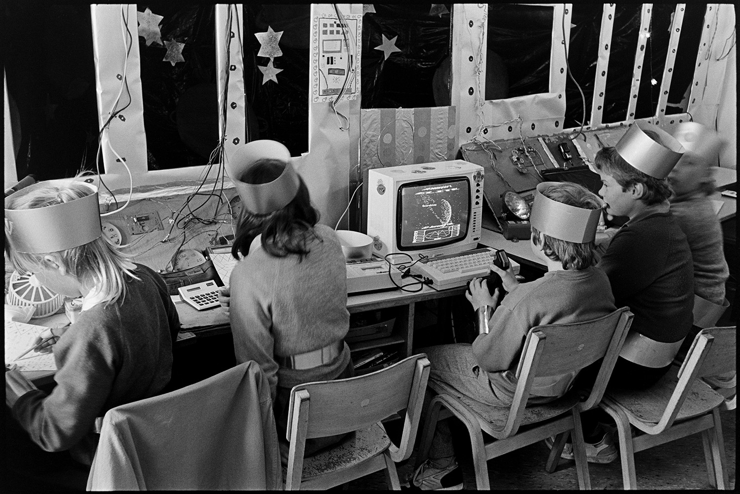 Schoolchildren working on space project pretending to be astronauts. 
[Boys and girls working on drawings for a space project in a classroom at Bideford Primary School. Two of the pupils are playing on a space themed computer game. They are all wearing hats.]