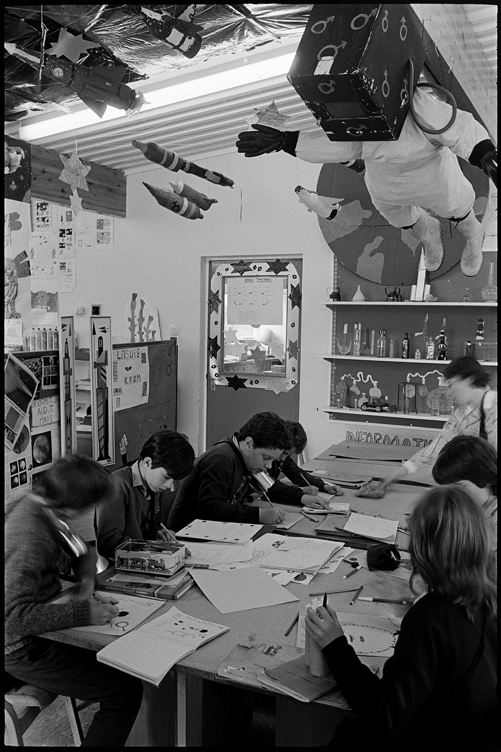 Schoolchildren working on space project pretending to be astronauts in spacecraft. 
[Boys and girls working at a desk in a classroom in Bideford Primary School, which is decorated with space themed objects, including rocket ships and an astronaut which are hung from the ceiling.