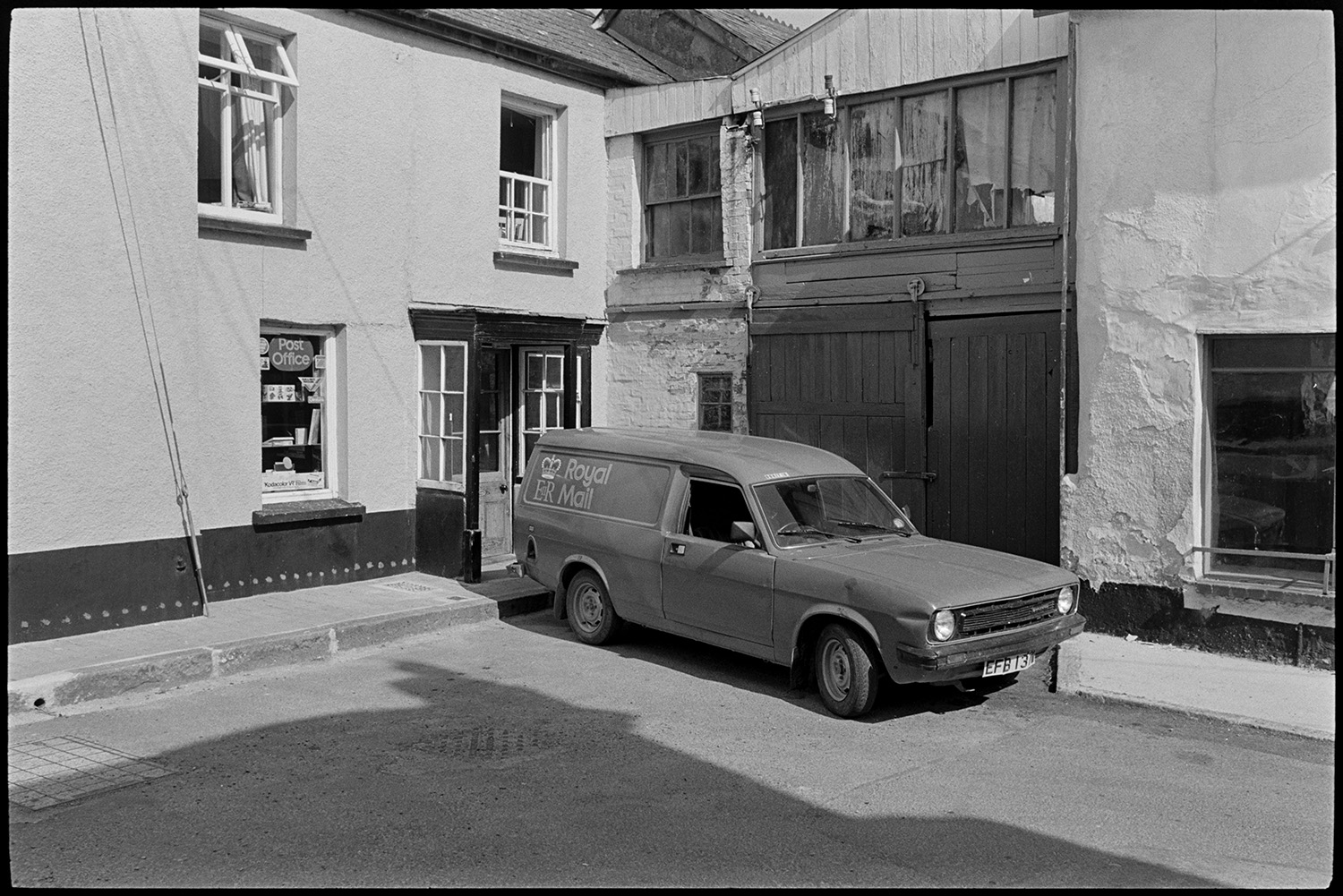 Street scene with Post Office. 
[A Royal Mail van parked outside Winkleigh Post Office and in front of a garage with a wooden door.]