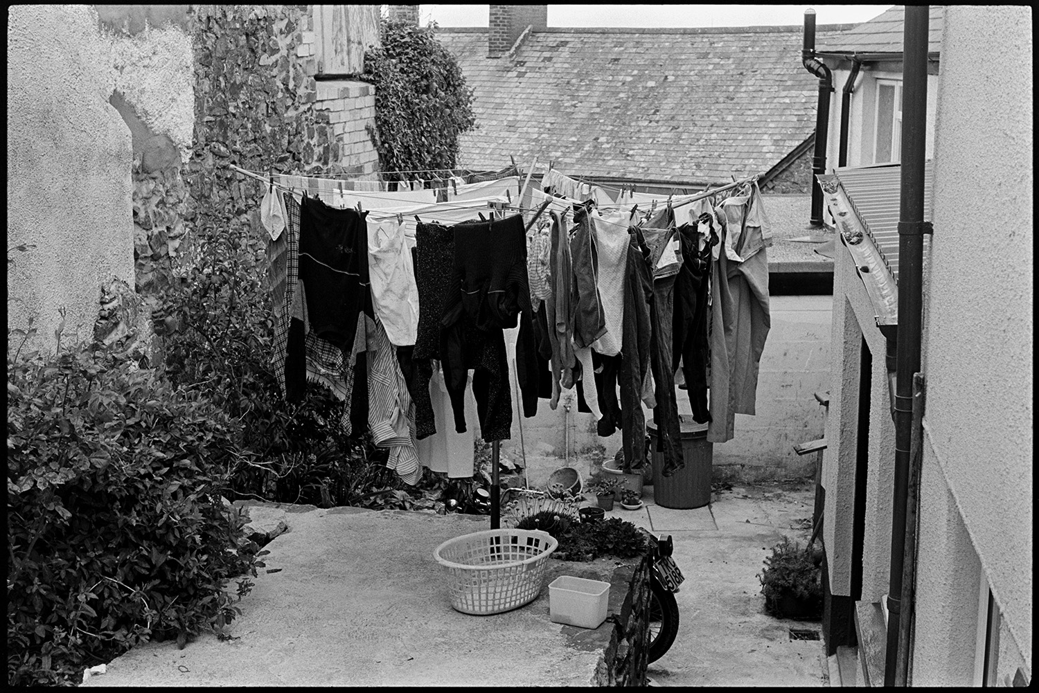 Washing line in yard. 
[Washing hung up to dry on a rotary washing line in the back yard of a house in Winkleigh. A washing basket and tub of pegs is on the patio. The wheel of a motorbike and a dustbin can also be seen in the yard.]
