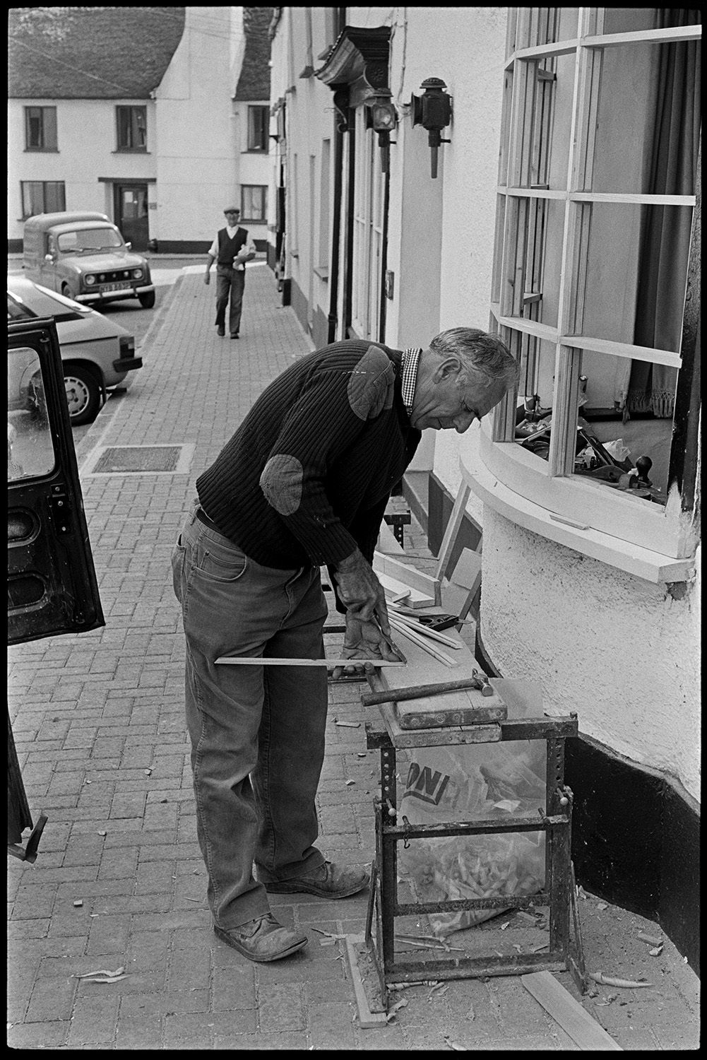 Man mending widow in village street. 
[A man fixing the window frame of a house in Winkleigh. He has a work bench set up on the pavement in front of the bay window. Another man is walking down the street towards him.]