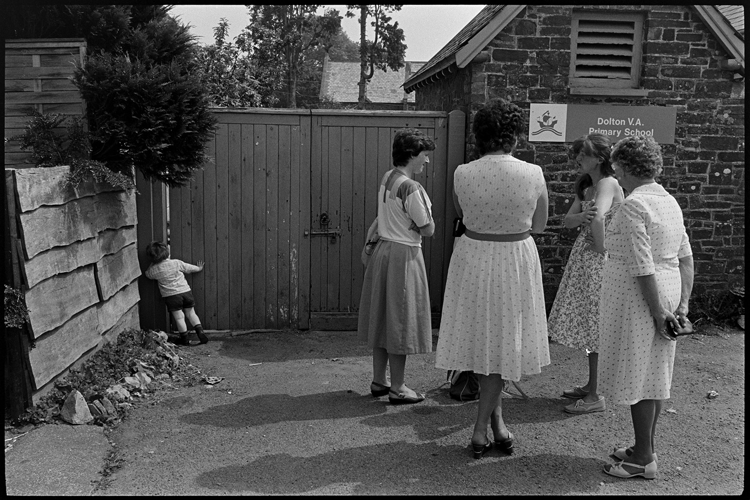 Women, mothers and children after school, in summer dresses. 
[Five women waiting for their children to finish school outside the gates of Dolton Primary School. They are wearing summer dresses. A young boy is peering through a gap by the side of the school gates in the background.]